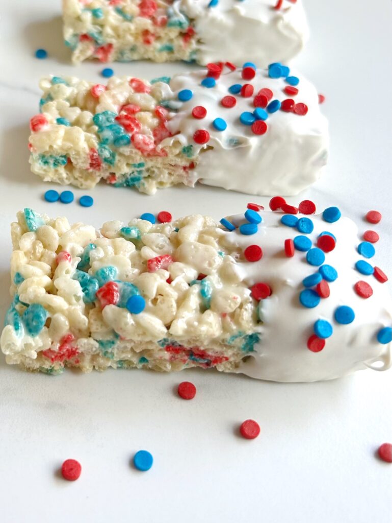 Three Rice Krispie treats lined up, each partially dipped in white candy melt and decorated with red and blue sprinkles
