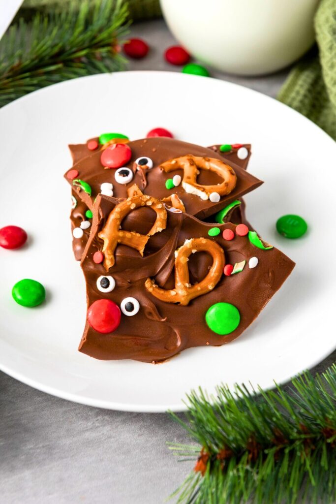 Close-up of chocolate bark pieces on a plate, each decorated like a reindeer with mini pretzels as antlers, candy eyes, and red and green candies for noses and accents