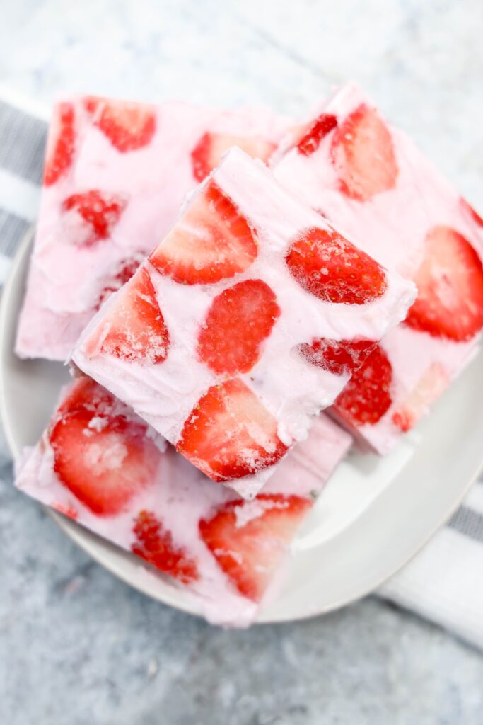 Close-up of square pieces of frozen yogurt bark with visible swirls of creamy pink yogurt and sliced strawberries, stacked on a white plate with a striped dish towel in the background
