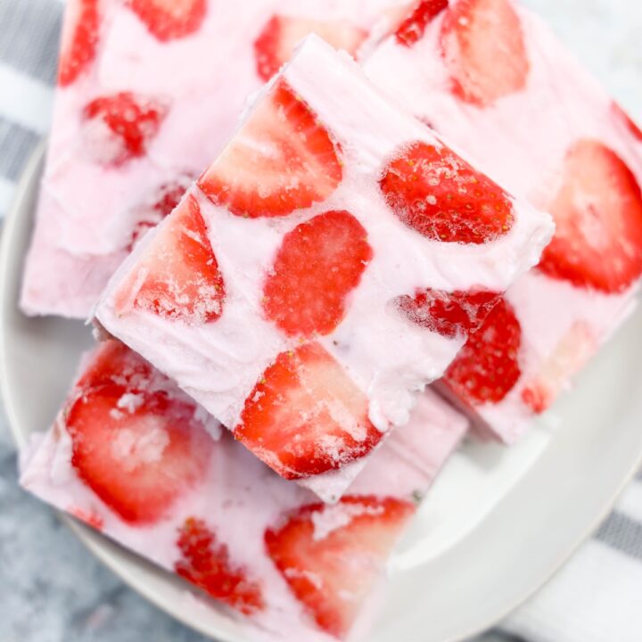 Close-up of square pieces of frozen yogurt bark with visible swirls of creamy pink yogurt and sliced strawberries, stacked on a white plate with a striped dish towel in the background