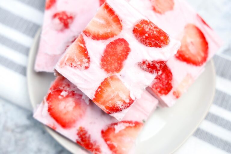 Close-up of square pieces of frozen yogurt bark with visible swirls of creamy pink yogurt and sliced strawberries, stacked on a white plate with a striped dish towel in the background