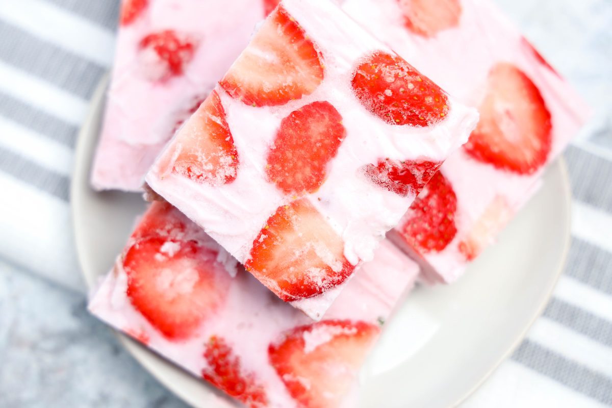 Close-up of square pieces of frozen yogurt bark with visible swirls of creamy pink yogurt and sliced strawberries, stacked on a white plate with a striped dish towel in the background