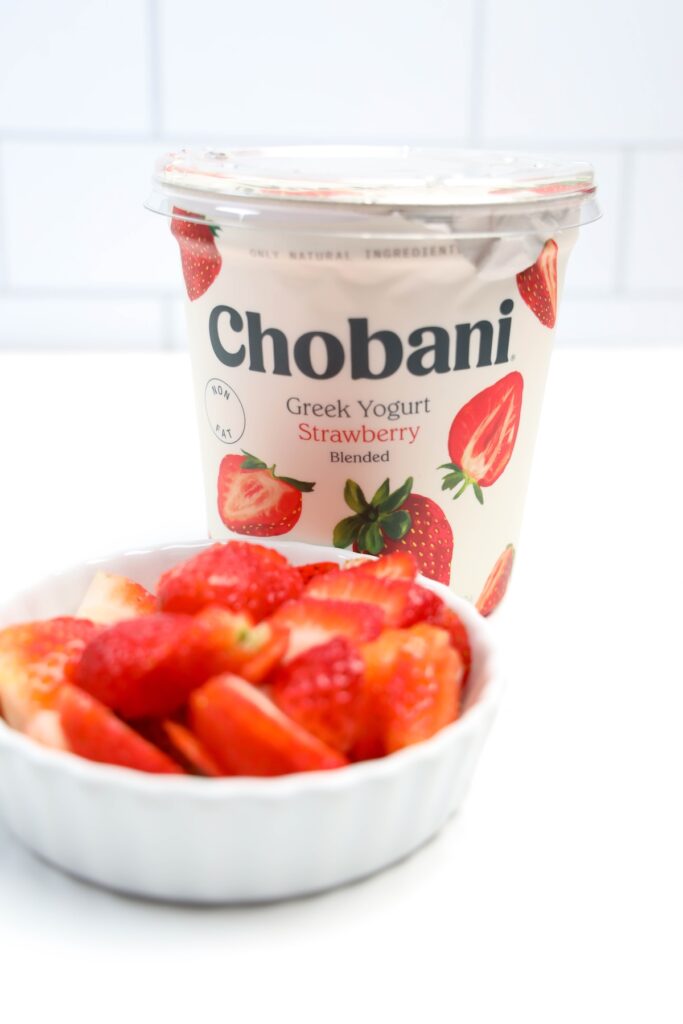 Overhead shot of ingredients for strawberry frozen yogurt bark, including a bowl of plain yogurt, fresh sliced strawberries, a small bowl of honey, and a few sprigs of mint arranged on a white surface