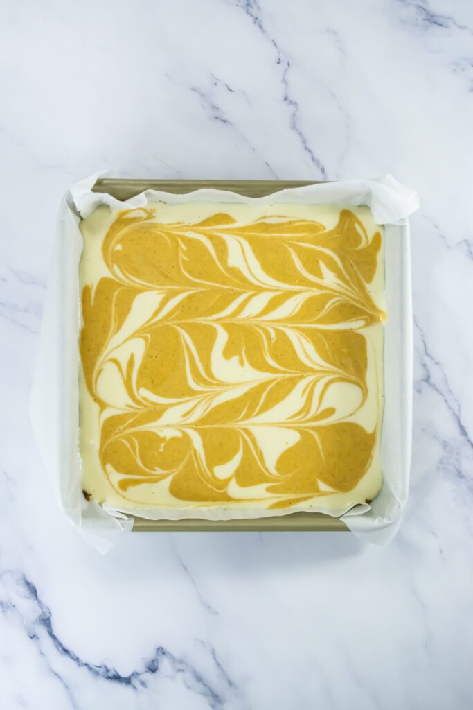 pumpkin cheesecake bar filling in a pan with regular cheesecake and pumpkin cheesecake swirled together