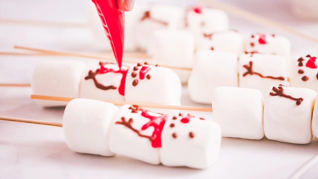 marshmallows kebabs with frosting being piped on to look like snowmen